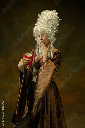 Astonished eating fried potato. Portrait of medieval young woman in vintage clothing on dark background. Female model as a duchess, royal person. Concept of comparison of eras, modern, fashion, beauty