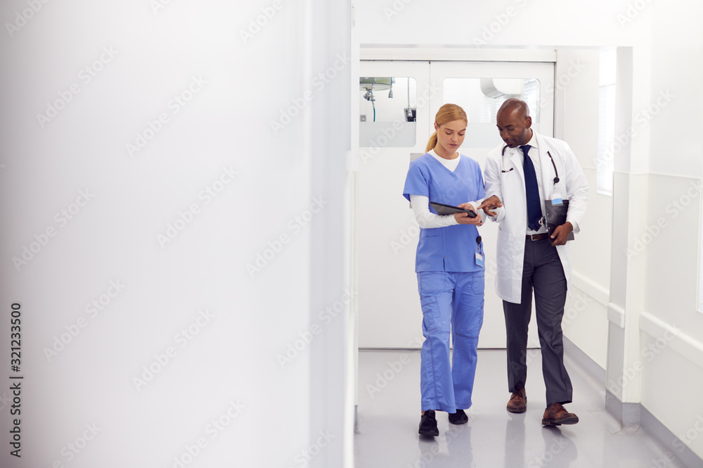 Doctor In White Coat And Nurse In Scrubs Looking At Digital Tablet In Hospital Corridor