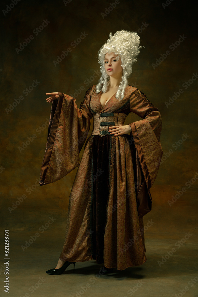 Elegance posing. Portrait of medieval young woman in brown vintage clothing on dark background. Female model as a duchess, royal person. Concept of comparison of eras, modern, fashion, beauty. Elegance posing. Portrait of medieval young woman in brown vintage clothing on dark background. Female model as a duchess, royal person. Concept of comparison of eras, modern, fashion, beauty.
