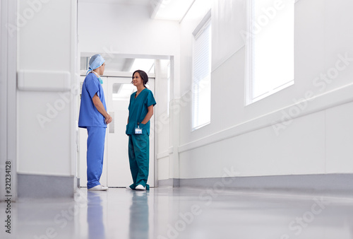 Male And Female Doctors Wearing Scrubs Meeting In Busy Hospital Corridor