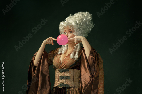 Pink bubble gum. Portrait of medieval young woman in brown vintage clothing on dark background. Female model as a duchess, royal person. Concept of comparison of eras, modern, fashion, beauty. photo