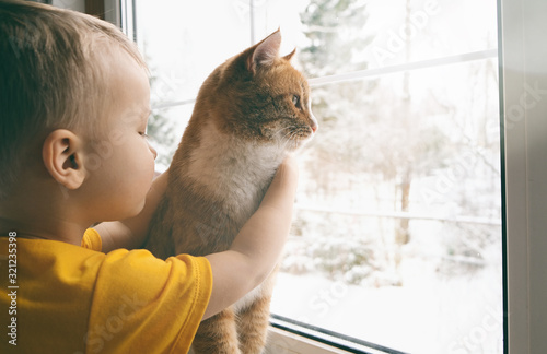 Child and pet at home. Red ginger tabby cat and little toddler boy looking out fo window watching falling snow. Domestic animal and kid friendship. Cozy scene, lifestyle. Copy space. photo