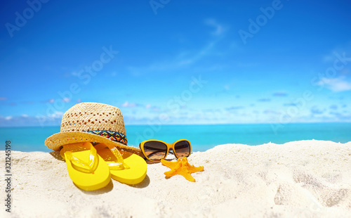 Fototapeta Naklejka Na Ścianę i Meble -  Concept summer beach holiday. Beach accessories - straw hat, glasses, starfish, yellow flip-flops on sandy tropical beach against blue sky with clouds on bright sunny day. Beautiful colorful image.