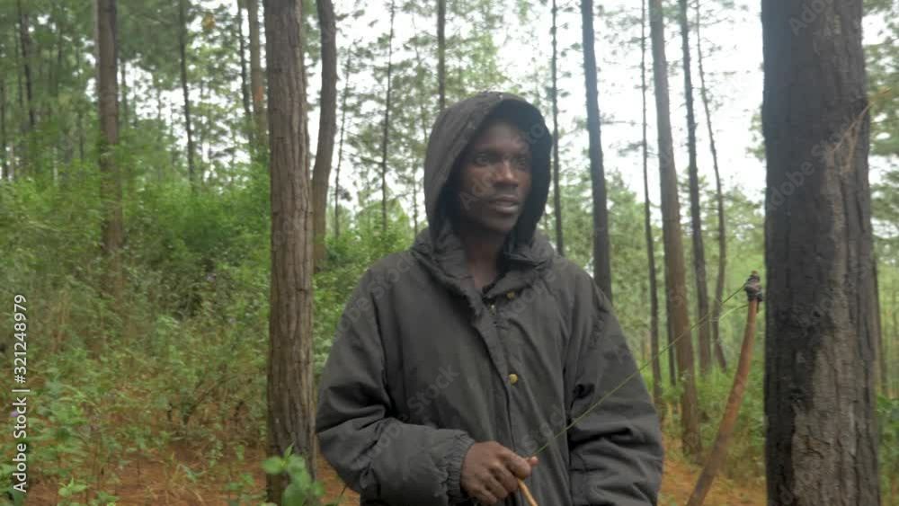 A slow motion shot tracking from in front an African hunter wearing a hooded coat as he walks through a forest hunting with a bow and arrow.