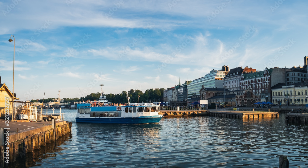 Fototapeta premium Tourist ship in Old Port of Helsinki, Finland, boat with tourists arrives ashore. Scenic summer panorama of Port pier architecture in Old Town under blue cloudy sky