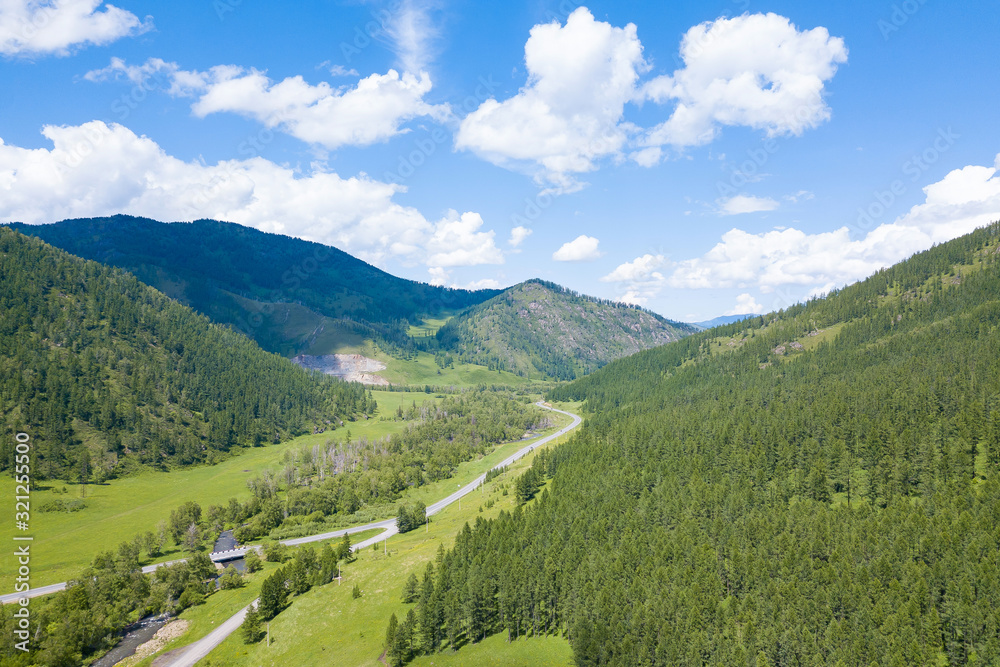 Aerial landscape with mountains, green trees, field, road and river under blue sky and clouds in summer