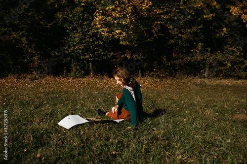 Portrait of young female artist sitting and working on painting outdoors, in the autumn landscape. She is in front of the canvas and drawing.She holds oil paints, artist brushes, canvas and palette. 