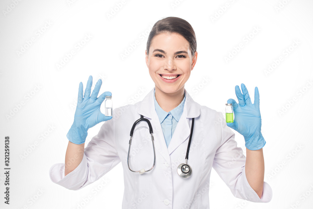 Beautiful doctor smiling at camera while holding jars with vaccines isolated on white