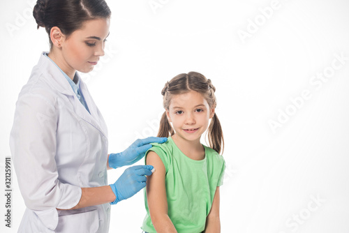 Female pediatrician with cotton wool rubbing shoulder of smiling kid isolated on white