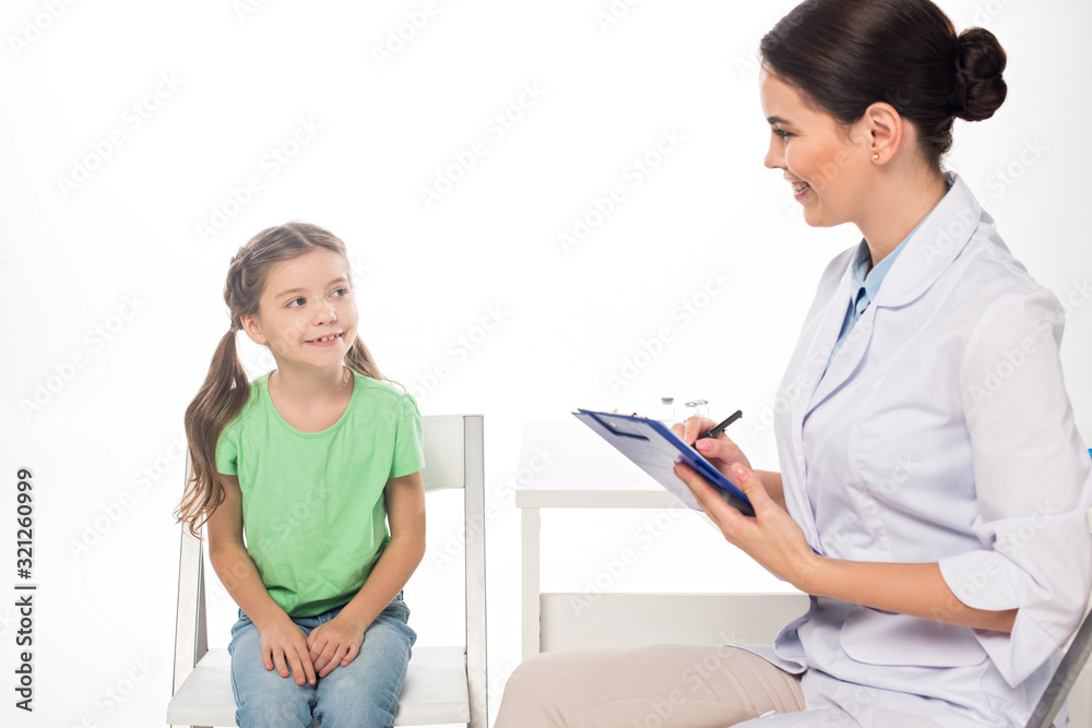 Smiling pediatrician with clipboard looking at kid on chair isolated on white Smiling pediatrician with clipboard looking at kid on chair isolated on white