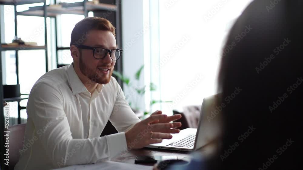 Handsome male manager with glasses and beard is consulting client, explaining deal details convincing buy services in modern office meeting against large window. Shooting in slow motion. Handsome male manager with glasses and beard is consulting client, explaining deal details convincing buy services in modern office meeting against large window. Shooting in slow motion.