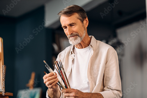 Portrait of senior painter holds a bunch of brushes choosing a one