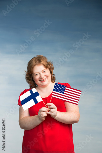 Red-haired plump woman holding Finlad and American flags in front of her on a blue background. photo