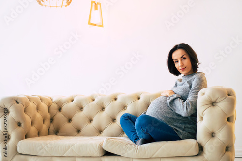 Future mom. Young pregnant mom in casual clothes is sitting on a couch at home in a lotus position and looking in the camera.