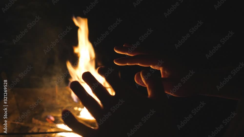 Male tourist warms his arms by a fire from metal brazier against the black background. Young man rubs his hands in front of an open bonfire from mangal at night. Warmth and tranquil concept. Top view