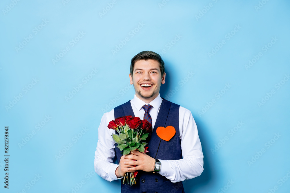 portrait of young surprised caucasian guy in stylish tuxedo, funny male stand surprisingly looking at camera, hold bouquet of beautiful red roses in hands. isolated blue background portrait of young surprised caucasian guy in stylish tuxedo, funny male stand surprisingly looking at camera, hold bouquet of beautiful red roses in hands. isolated blue background