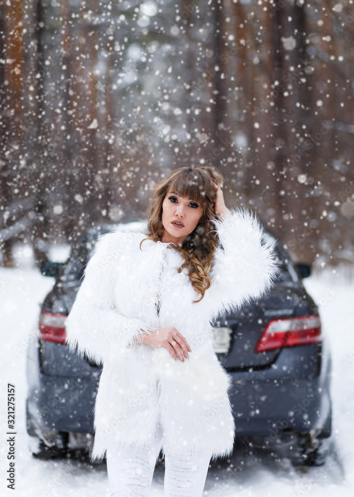A beautiful pretty cheerful funny young girl with curly hair and a snow white smile in a white fur coat and a warm hat looks out of the car window against the background of a winter forest A beautiful pretty cheerful funny young girl with curly hair and a snow white smile in a white fur coat and a warm hat looks out of the car window against the background of a winter forest