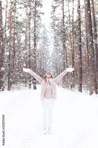 A beautiful pretty young girl with curly hair and a snow-white smile in a pink fur coat is walking and fooling around in the winter forest against the background of snow and trees, enjoy