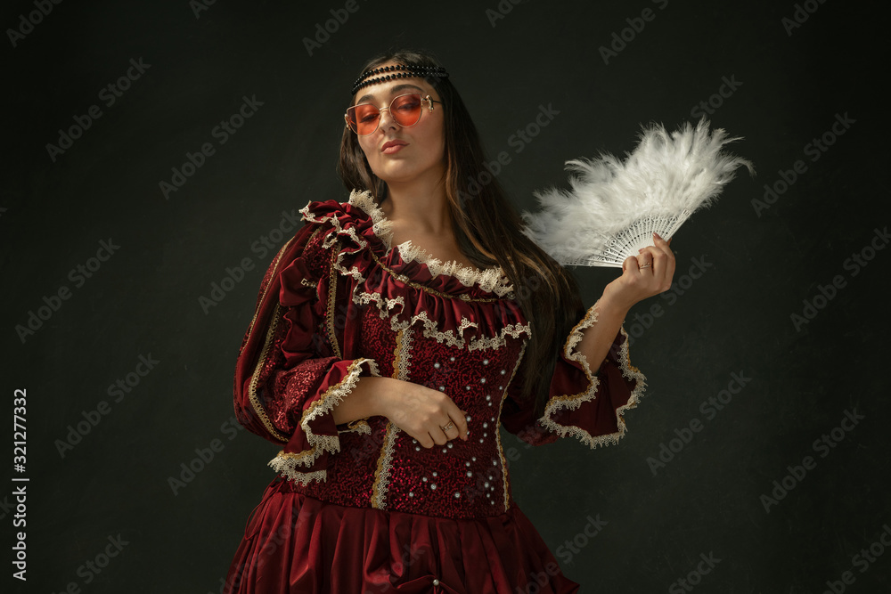 Red eyewear and fluffy fan. Portrait of medieval young woman in red vintage clothing on dark background. Female model as a duchess, royal person. Concept of comparison of eras, modern, fashion, beauty