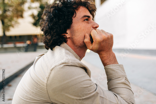 Side view of young man with curly hair looking at one side, sitting outside. Portrait of handsome male with curly hair has pensive expression, take a rest in the city street. photo