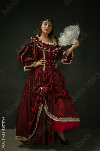Portrait of medieval young woman in red vintage clothing posing with fluffy fur on dark background. Female model as a duchess, royal person. Concept of comparison of eras, modern, fashion, beauty.