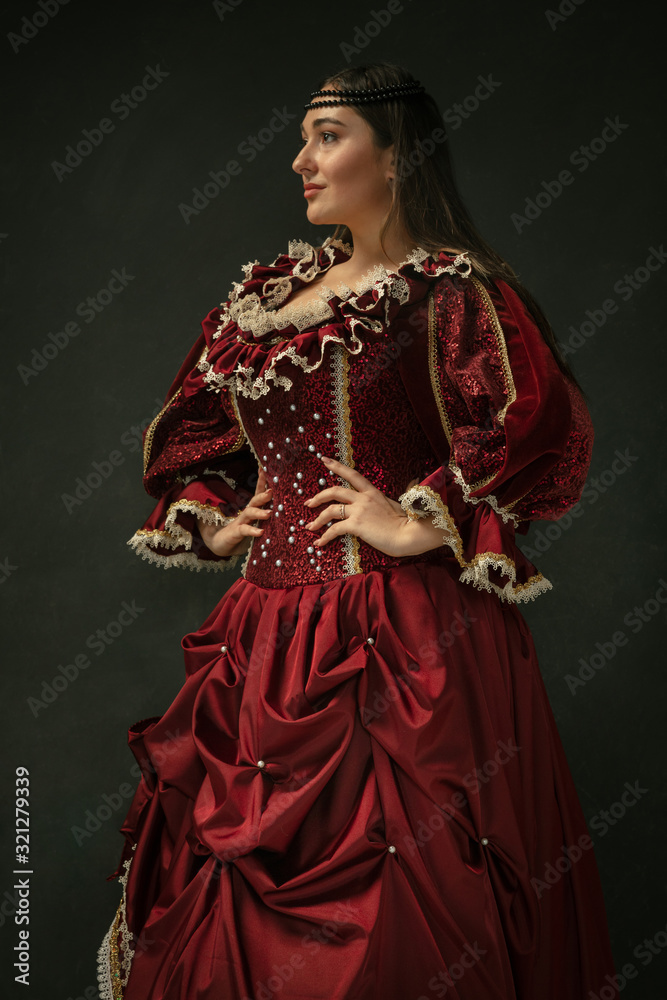 Posing as royal. Portrait of medieval young woman in red vintage clothing standing on dark background. Female model as a duchess, royal person. Concept of comparison of eras, modern, fashion, beauty.