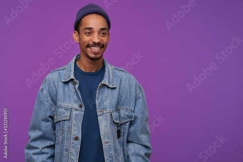 Cheerful young attractive bearded brunette man with dark skin looking positively aside with charming smile, wearing blue cap, pullover and jeans coat while posing over purple background