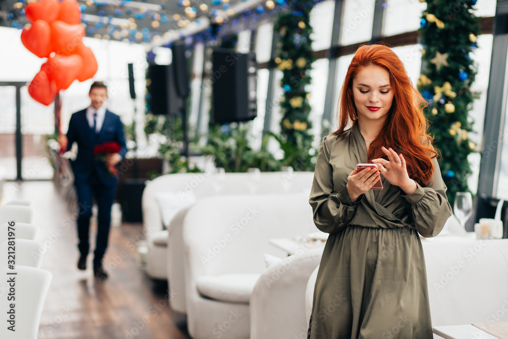 attractive redhaired lady wait for a man sweetheart in executive restaurant, beautiful woman using smartphone while waiting, celebration of st valentines day