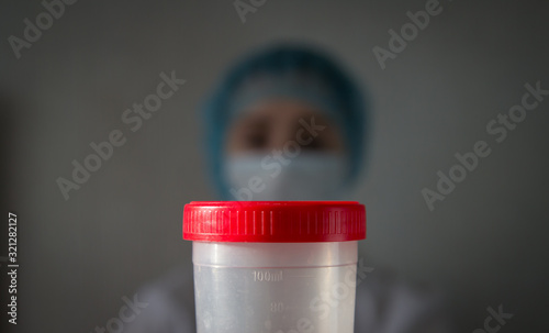  A plastic container with a red lid for urine in the foreground and a medical nurse (paramedic) in a mask in the background.