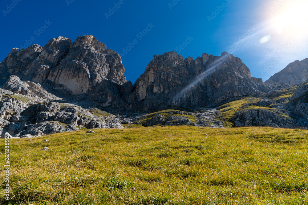 Obraz premium Sonne als Gegenlicht vor steiler Almwiese mit Dolomiten Berge im Hintergrund