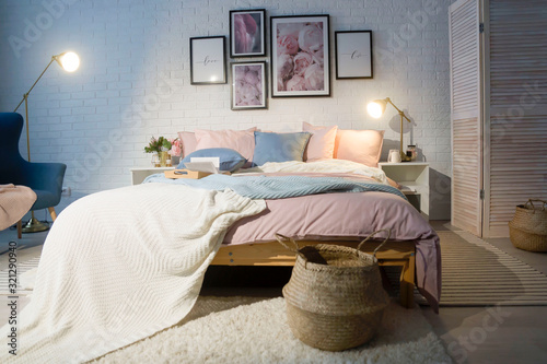 Cozy, feminine bedroom with pink bed, decorative cushions and plant on a wooden stool standing against white, empty wall. Real photo with a place for your furniture.