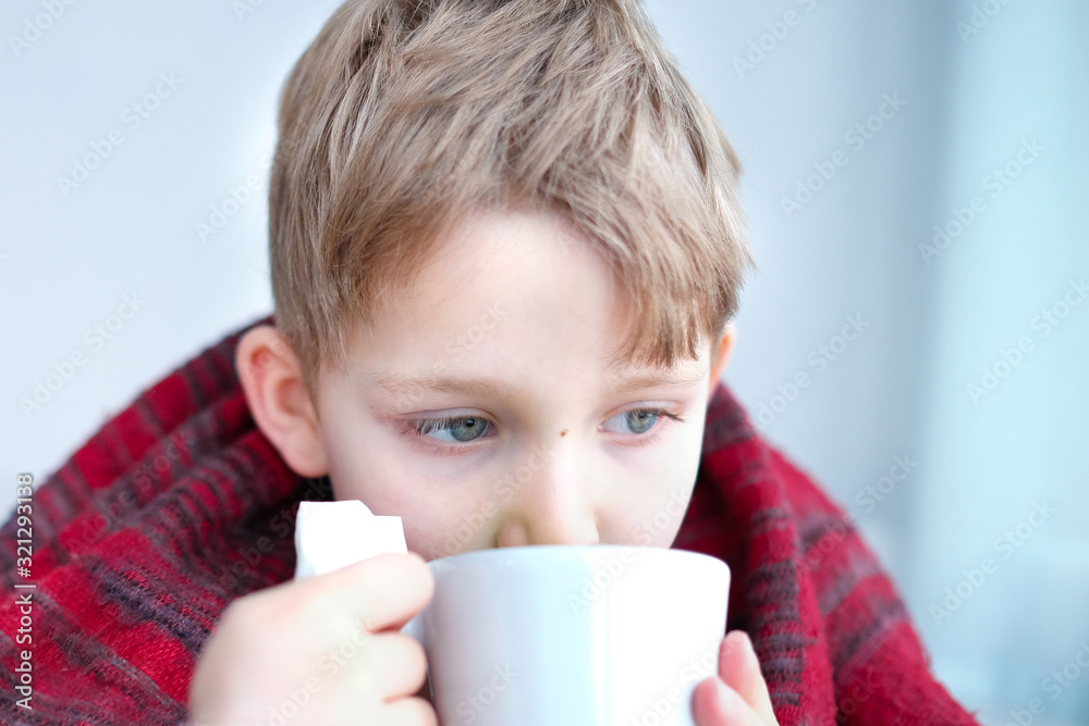 a small boy of the European race is ill, wipes his nose with a napkin and drinks a hot drink from a Cup, cover with a warm blanket in front of the window. concept of childhood diseases a small boy of the European race is ill, wipes his nose with a napkin and drinks a hot drink from a Cup, cover with a warm blanket in front of the window. concept of childhood diseases