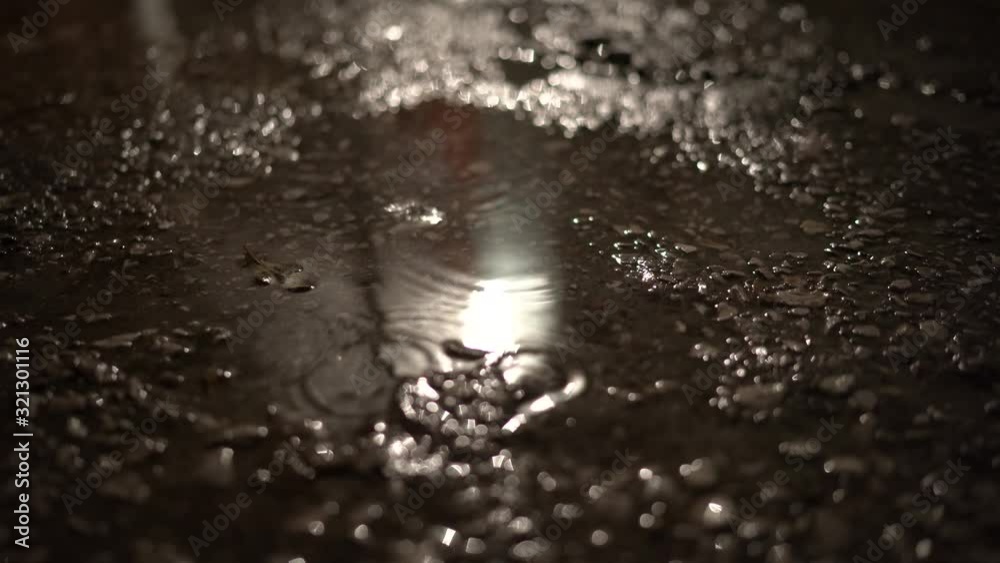 Rain drops in puddle on pavement at night street light reflected in water.