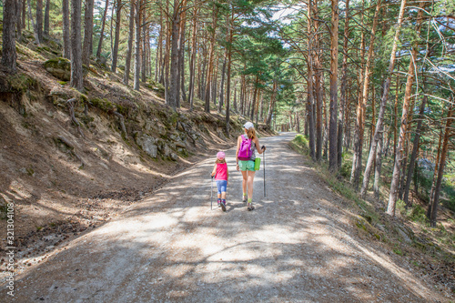 from behind mother and daughter, five years old girl, with caps and trekking sticks, hiking on track in forest of Canencia mountain (Madrid, Spain, Europe) photo