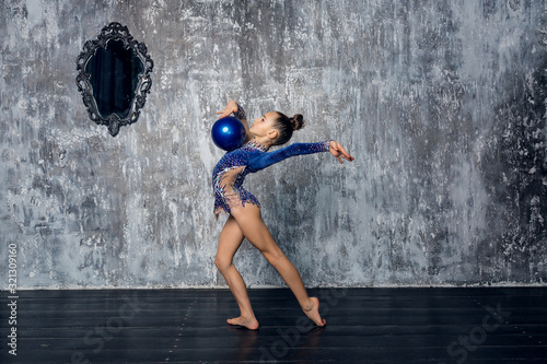 Girl gymnast in a blue suit makes exercise with a ball against a gray wall.