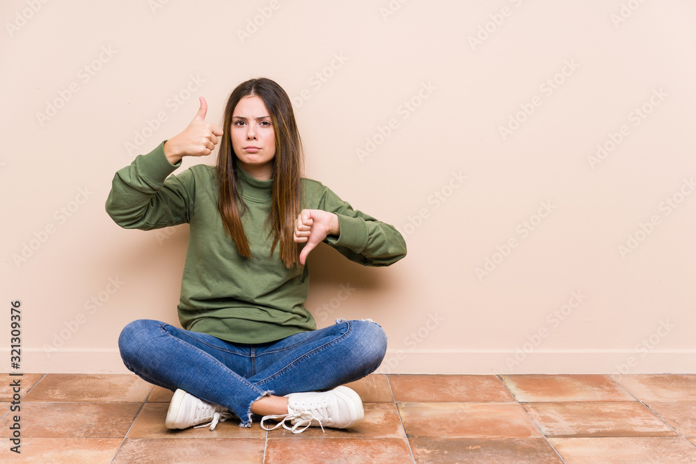Young caucasian woman sitting on the floor isolated showing thumbs up and thumbs down, difficult choose concept Young caucasian woman sitting on the floor isolated showing thumbs up and thumbs down, difficult choose concept