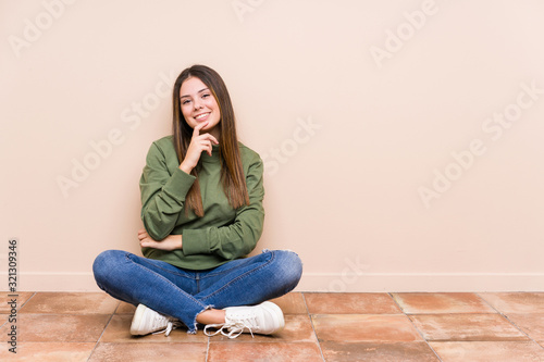 Young caucasian woman sitting on the floor isolated smiling happy and confident, touching chin with hand.