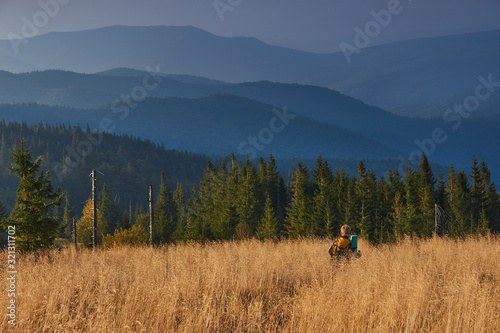 Hiker is walking along the trail through tall grass in the mountains. View on the wooded hills and hazy peaks in the distance at sunset. Concept traveling and adventuring.