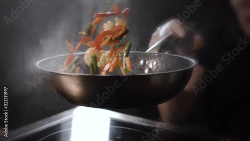Cook in transparent sterile gloves in the kitchen throws up a hot frying wok with chicken, vegetables and noodles in a black pan