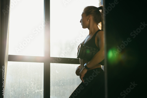 beautiful young girl trainer stands near the window and holds in her hands a roll for warming up muscles in leggins. Loft gym in which the sun shines