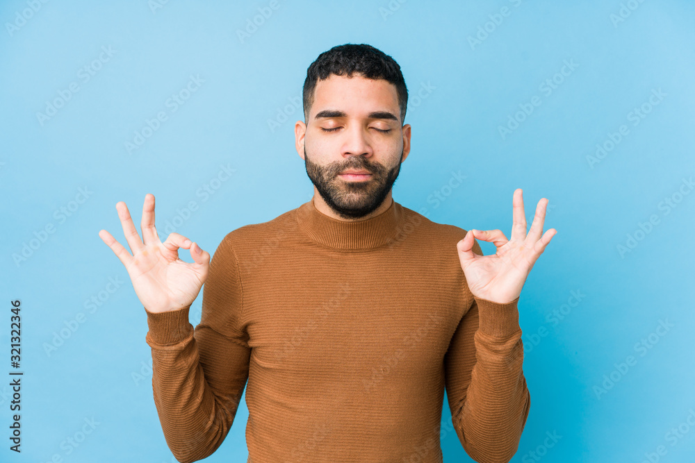 Fototapeta premium Young latin man against a blue background isolated relaxes after hard working day, she is performing yoga.