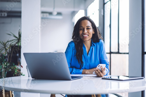 Happy ethnic businesswoman browsing smartphone while working on laptop at office table