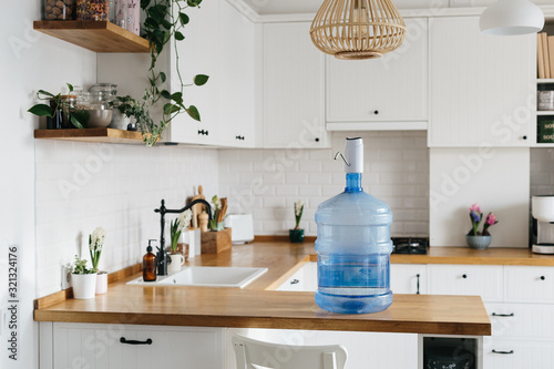 A larger bottle of clean water 19 liters with automatic white pomp in the interior of the apartment with a white kitchen in the background.