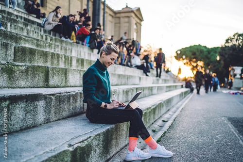 Side view of talented happy hipster girl with electronic stylus in hand resting at city urban area and using public internet connection for online messaging in group chat for creative artists