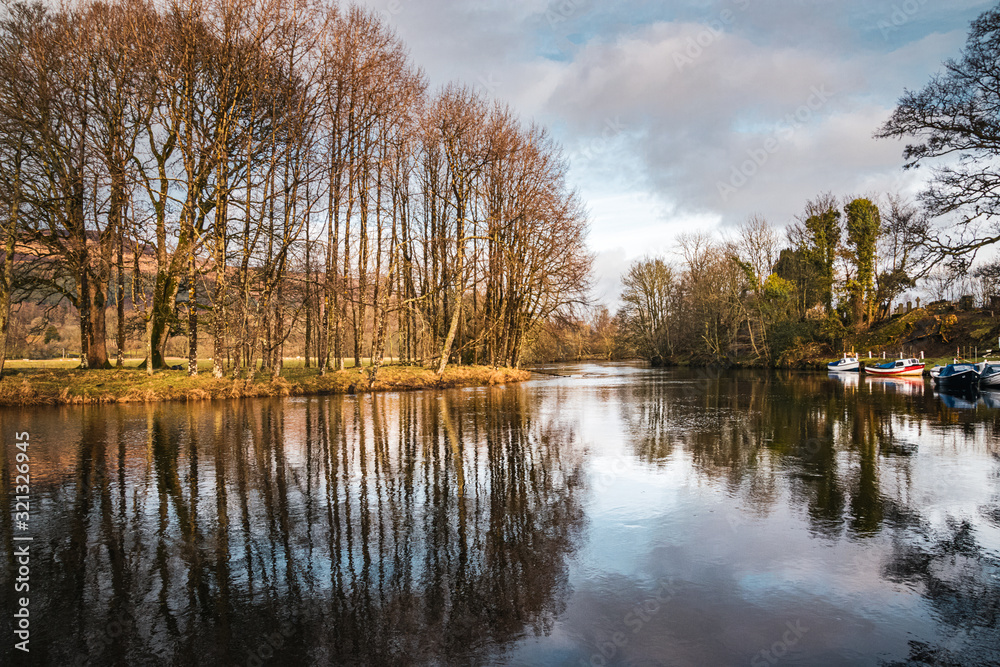 Trees reflecting in the still surface of River Lochay in Killin, on a winter afternoon, with a few boats moored at the local jetty. Perthshite Scotland.