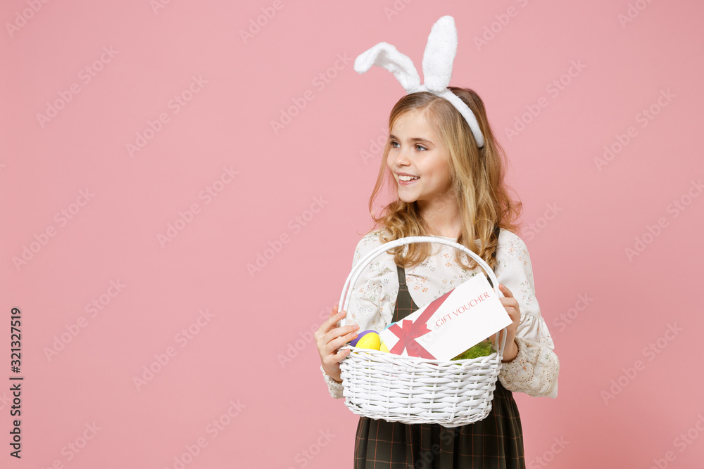 Little pretty blonde kid girl 11-12 years old in spring dress, bunny rabbit ears on head hold in hand wicker basket colorful eggs gift coupon isolated on pastel pink background. Happy Easter concept.