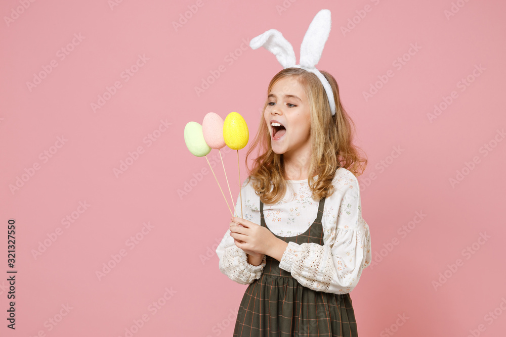 Little pretty blonde kid girl 11-12 years old in spring dress, bunny rabbit ears hold in hand carries dyed eggs on sticks have fun celebrate isolated on pastel pink background. Happy Easter concept.