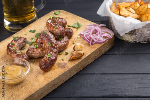 Grilled sausages from a BBQ served with a mustard, fried potatoes, garlic, herbs, beer on a wooden board all served on a black wooden table. Delicious german sausages Perfect for Octoberfest.