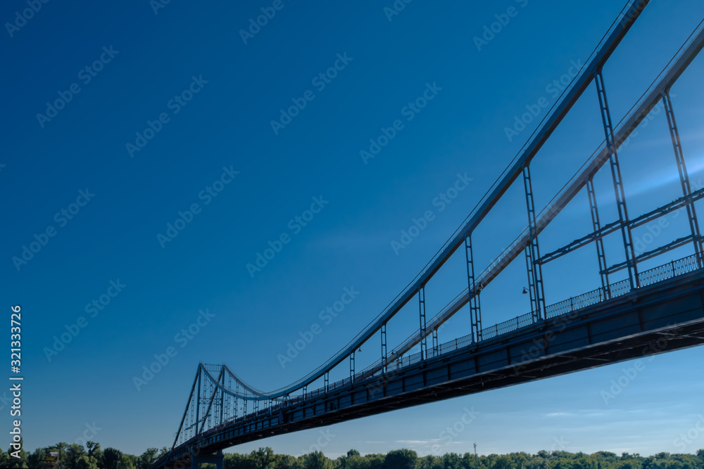 View of the long beautiful bridge from the side below. A pathway to the other side with trees against a blue sky without clouds on a clear day. Minimalist concept of joining with a place for text.