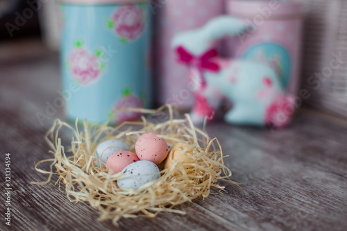 Pastel colored easter eggs in a nest on a wooden table. Against the background of handmade textile rabbit and tin cans for loose in pink and blue colors. Foreground focus